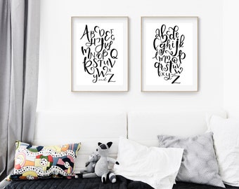 May include: Two framed art prints featuring black calligraphy alphabets on white backgrounds. The prints are in gold frames and are displayed above a white couch with decorative pillows and stuffed animals. The letters are in a cursive style.