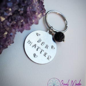 以下が含まれることがあります： シルバーのキーホルダーに、ハートが両側に付いた「You Matter」と書かれた丸いチャームが付いています。チャームは、黒い溶岩ビーズが付いたシルバーのキーリングに付いています。