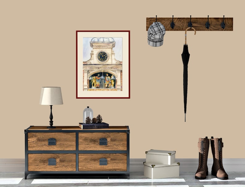 May include: A framed watercolor painting of a clock tower with a clock face and figures on the side. The painting is hanging on a beige wall with a wooden coat rack with a hat and umbrella hanging on it. A wooden dresser with four drawers is in the foreground.