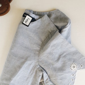 May include: A light blue cotton shirt with a white lace trim on the sleeves. The shirt has a label that says "Abby" on it.