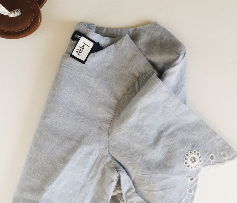 May include: A light blue cotton shirt with a white lace trim on the sleeves. The shirt has a label that says "Abby" on it.