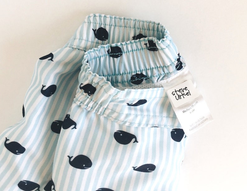 May include: Blue and white striped swim trunks with a whale pattern. The trunks have an elastic waistband and a label that reads "Steve Urkel" with the text "Medium/Medium" and "4-5 yrs" underneath.