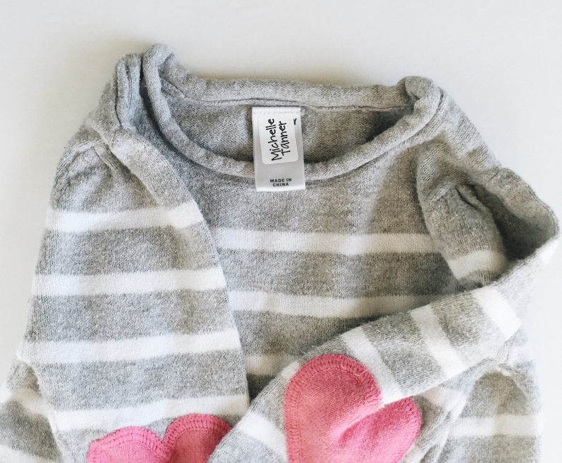 May include: Gray and white striped sweater with pink heart patches on the elbows. The sweater has a tag that reads "Michelle  Turner" and "Made in China".