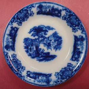 May include: A round ceramic saucer featuring a white base with a blue transferware pattern. The center displays a landscape with trees and buildings, while the rim is decorated with a repeating floral and architectural scene in deep blue.
