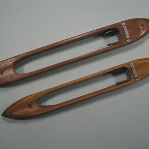 Op de afbeelding: Twee langwerpige houten weefspoelen met gladde, gepolijste bruine oppervlakken. Elke spoel heeft een centrale open gleuf, een puntig uiteinde aan beide kanten en een klein metalen spanningsmechanisme met een gekartelde rand in het hout.