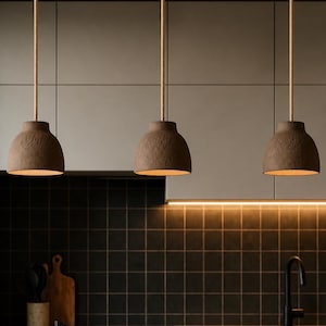 May include: Three pendant lights with textured brown shades hang above a kitchen island. The kitchen features a black tiled splashback, a bowl of oranges and lemons, and a black tap. The lights are suspended by rope cords.