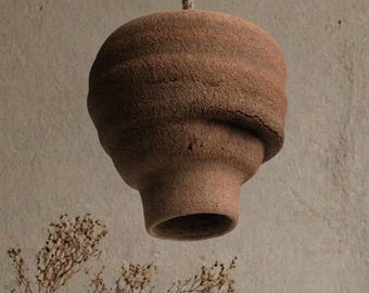 May include: A brown, textured, hanging light fitting with an unusual, organic shape. The light hangs from a rope above a wooden shelf with books, a vase, and a bowl. The room has a neutral colour palette.
