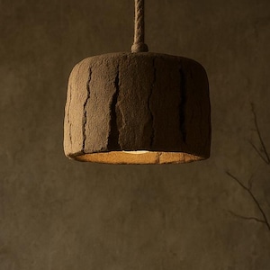 May include: A brown, textured pendant light hangs from a rope. The cylindrical lampshade has a rough, bark-like appearance. The warm light from within illuminates the textured surface. The image has a rustic, earthy aesthetic.