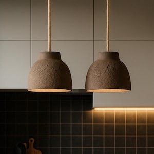 May include: Two brown textured pendant lights hang from beige cords above a kitchen island. A bowl of oranges and lemons sits on the countertop. The kitchen has a black tiled backsplash and a black tap. The lights are a feature of the kitchen.