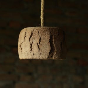 May include: A hanging, textured, brown pendant light fixture. The light has a rectangular shape with a rough, cracked surface. A natural rope suspends the light from above. The background is a blurred brick wall.