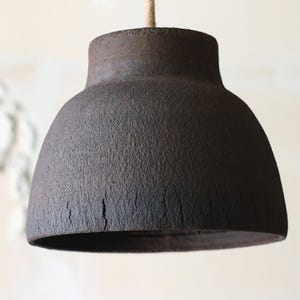 K&ouml;nnte beinhalten: Eine dunkelbraune, strukturierte Pendelleuchte mit einem glockenf&ouml;rmigen Schirm. Das Licht h&auml;ngt an einem Naturseil. Der Schirm hat eine raue, unebene Oberfl&auml;che und einen leicht ausgestellten Sockel.