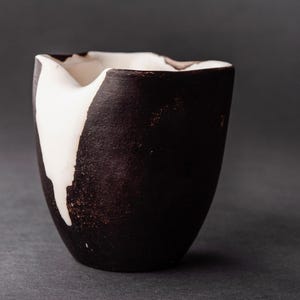 Könnte beinhalten: Eine schwarze und weiße Keramikvase mit unregelmäßiger, asymmetrischer Form. Die Vase hat eine weiße Glasur, die an der Seite herunterläuft und ein einzigartiges Design erzeugt.