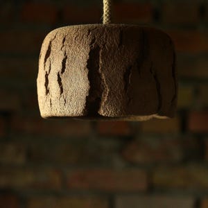 May include: A brown, textured, cylindrical pendant light fixture hanging from a rope. The light has a rough, uneven surface with vertical grooves, and is set against a blurred brick wall background. The fixture is illuminated from below.
