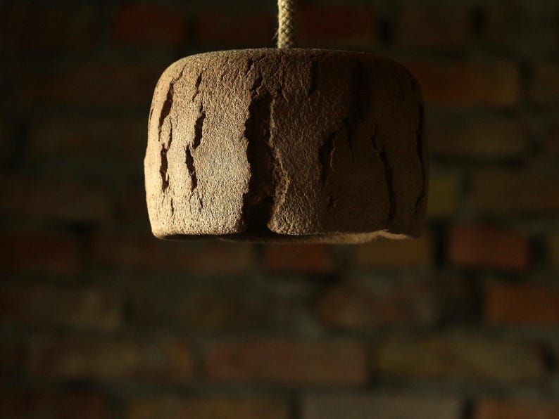May include: A brown, textured, cylindrical pendant light fixture hanging from a rope. The light has a rough, uneven surface with vertical grooves, and is set against a blurred brick wall background. The fixture is illuminated from below.