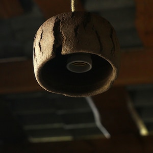 May include: A hanging pendant light fixture with a textured, brown, rock-like shade. The light fixture has a wide opening at the bottom, revealing the light bulb socket. The fixture is suspended by a rope.