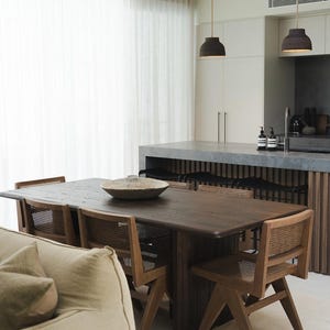 K&ouml;nnte beinhalten: Ein Esstisch aus dunklem Holz mit sechs passenden St&uuml;hlen. Die St&uuml;hle haben geflochtene R&uuml;ckenlehnen und Holzrahmen. Eine gro&szlig;e Holzschale steht in der Mitte des Tisches. Der Raum hat eine neutrale Farbpalette mit wei&szlig;en Vorh&auml;ngen und Pendelleuchten.