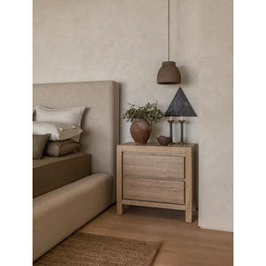 K&ouml;nnte beinhalten: Schlafzimmerinterieur mit einem Nachttisch aus hellem Holz mit zwei Schubladen, auf dem sich eine Vase mit Gr&uuml;n, eine schwarze dreieckige Lampe und zwei Kerzenhalter befinden. Ein Bett mit beigem Kopfteil und Kissen ist ebenfalls sichtbar.