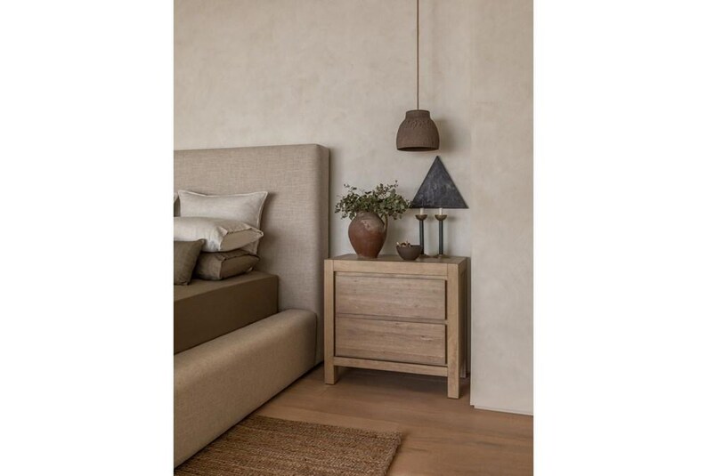 K&ouml;nnte beinhalten: Schlafzimmerinterieur mit einem Nachttisch aus hellem Holz mit zwei Schubladen, auf dem sich eine Vase mit Gr&uuml;n, eine schwarze dreieckige Lampe und zwei Kerzenhalter befinden. Ein Bett mit beigem Kopfteil und Kissen ist ebenfalls sichtbar.