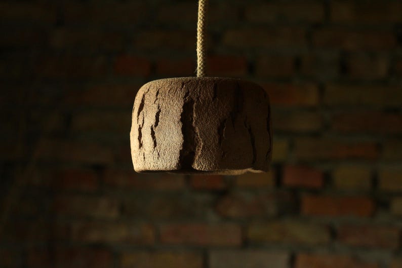 May include: A brown, textured pendant light fixture with a rough, bark-like appearance. The light hangs from a beige rope against a blurred brick wall background. The fixture is cylindrical with vertical grooves.