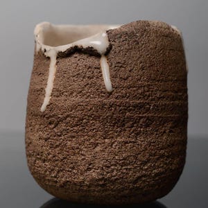 Può includere: Un vaso in ceramica fatto a mano, con un esterno marrone strutturato e un interno bianco. La glassa bianca cola lungo il lato, aggiungendo un dettaglio unico. Il vaso ha una base arrotondata e un bordo leggermente irregolare, che ne evidenzia la qualità artigianale.