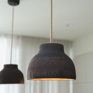 K&ouml;nnte beinhalten: Zwei dunkelbraune Pendelleuchten mit strukturierten Oberfl&auml;chen h&auml;ngen von der Decke. Die Leuchten haben ein glockenf&ouml;rmiges Design und sind an beigefarbenen Schn&uuml;ren aufgeh&auml;ngt. Das Innere der Leuchten leuchtet warm.