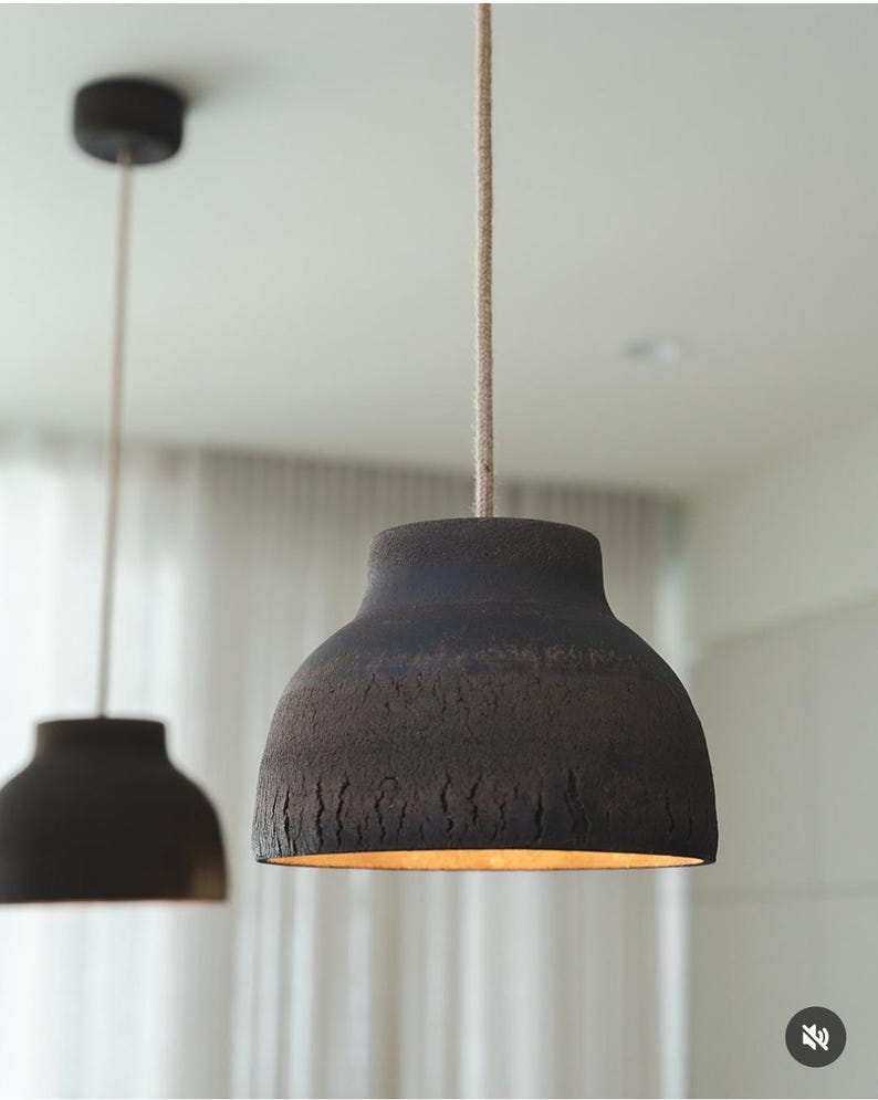 K&ouml;nnte beinhalten: Zwei dunkelbraune Pendelleuchten mit strukturierten Oberfl&auml;chen h&auml;ngen von der Decke. Die Leuchten haben ein glockenf&ouml;rmiges Design und sind an beigefarbenen Schn&uuml;ren aufgeh&auml;ngt. Das Innere der Leuchten leuchtet warm.