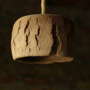May include: A handcrafted, brown ceramic pendant light fixture. The cylindrical shade has a textured, bark-like surface with vertical grooves. A natural rope suspends the light from above, creating a rustic aesthetic.