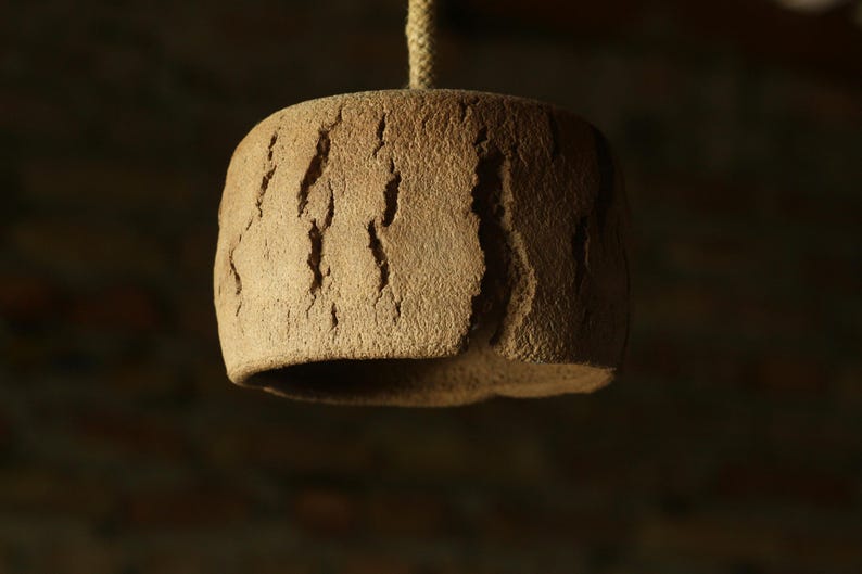 May include: A handcrafted, brown ceramic pendant light fixture. The cylindrical shade has a textured, bark-like surface with vertical grooves. A natural rope suspends the light from above, creating a rustic aesthetic.