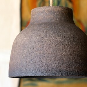 K&ouml;nnte beinhalten: Eine dunkelbraune, strukturierte Keramik-Pendelleuchte. Die Lampe hat einen abgerundeten, glockenf&ouml;rmigen Schirm und eine strukturierte Oberfl&auml;che. Das Licht h&auml;ngt an einem naturfarbenen Seil. Der Hintergrund ist unscharf.
