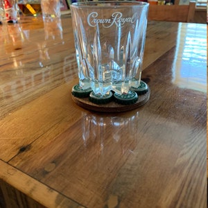 May include: A clear glass with the words "Crown Royal" etched on the side, sitting on a brown coaster made of bottle caps.