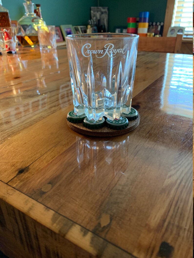 May include: A clear glass with the words "Crown Royal" etched on the side, sitting on a brown coaster made of bottle caps.