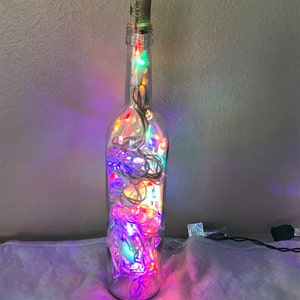 May include: A clear glass bottle with a cork stopper filled with colorful string lights. The lights are red, green, blue, and white. The bottle is sitting on a white fuzzy surface.