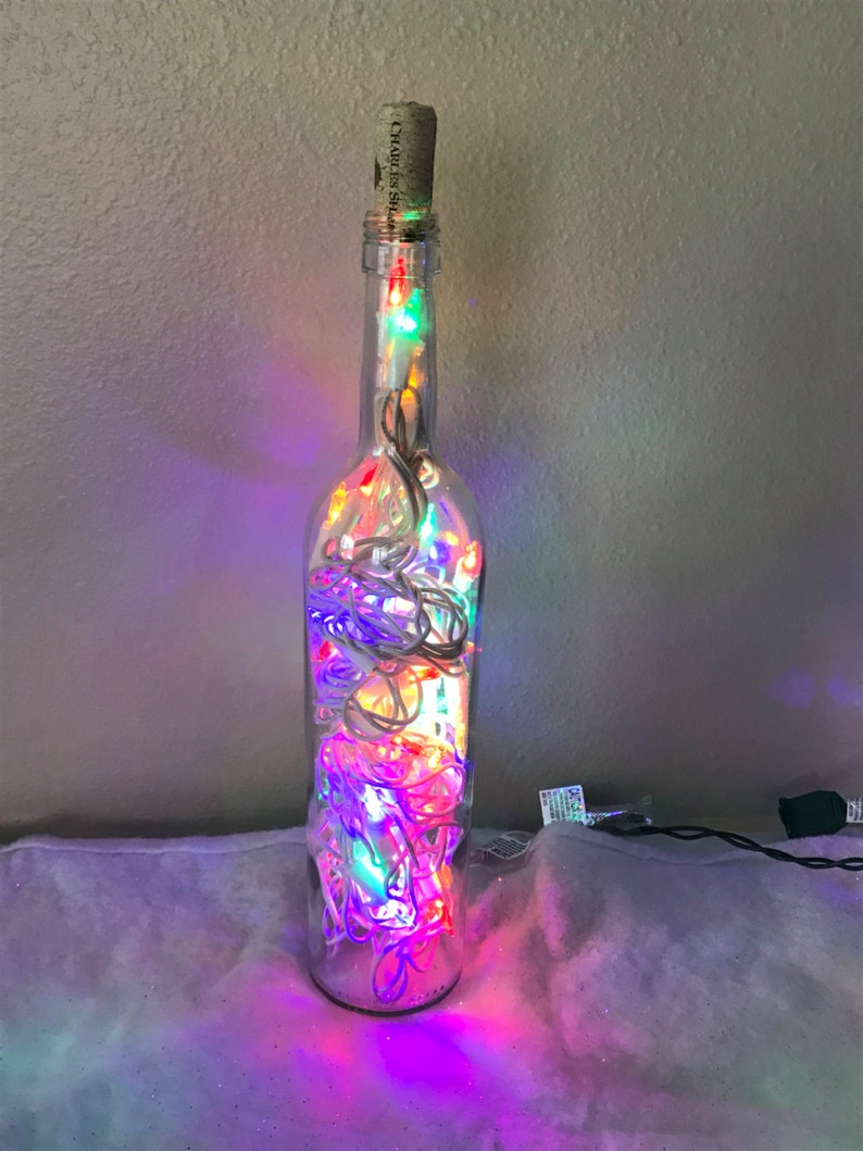 May include: A clear glass bottle with a cork stopper filled with colorful string lights. The lights are red, green, blue, and white. The bottle is sitting on a white fuzzy surface.