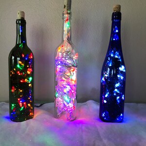 May include: Three glass bottles with different colored glass, each filled with white string lights. The bottles are sitting on a white surface.