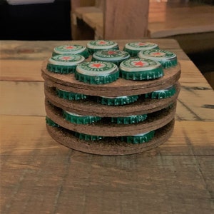 May include: A stack of brown cork coasters with green bottle caps on top. The bottle caps have a red star and the words "Original Quality" printed on them.
