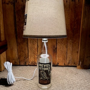 May include: A table lamp made from a clear glass bottle with a brown label that reads "Revel Stoke" and a beige fabric lampshade with a brown trim.