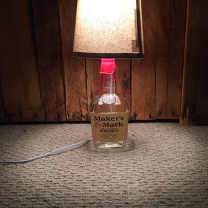 May include: A lamp made from a Maker's Mark whiskey bottle with a brown lampshade. The lamp is sitting on a light brown carpet.
