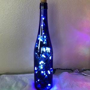 May include: A cobalt blue glass bottle with a cork top filled with white fairy lights. The bottle is lit up and casting a blue glow.