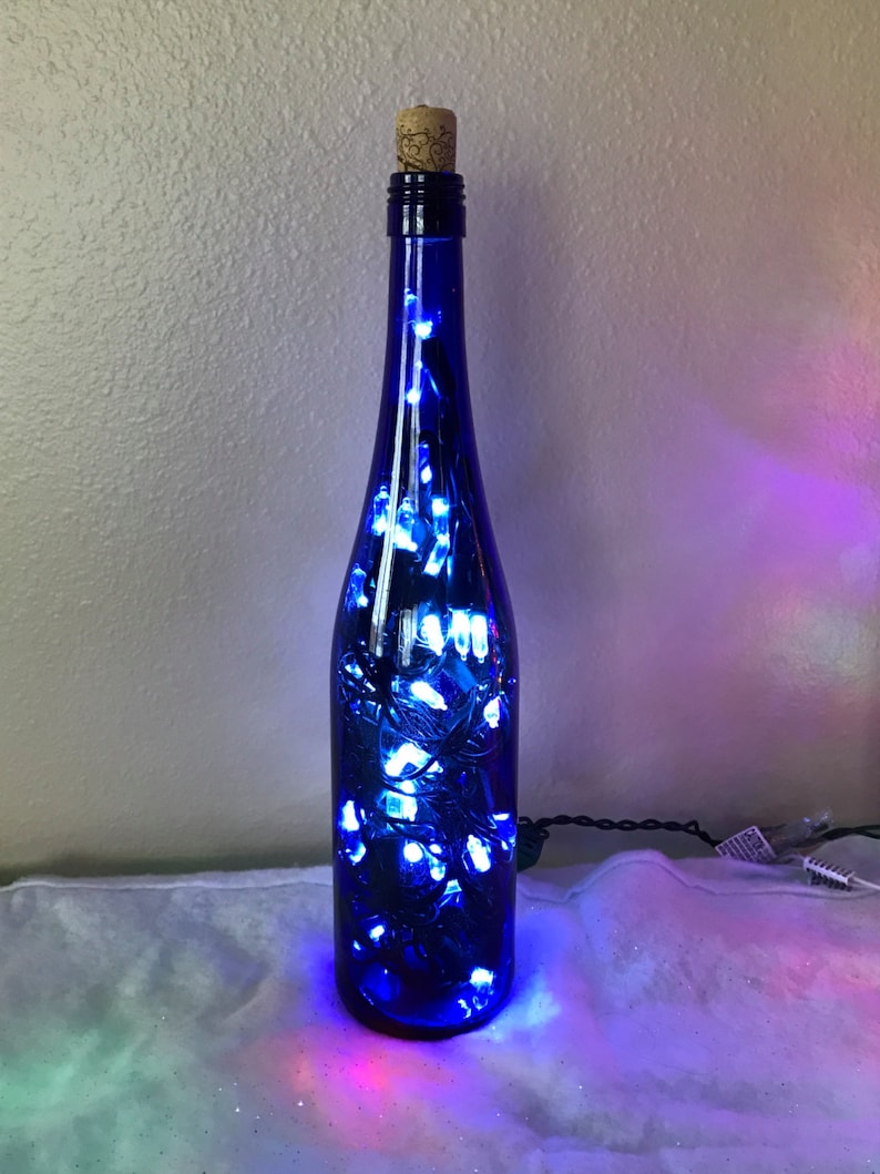 May include: A cobalt blue glass bottle with a cork top filled with white fairy lights. The bottle is lit up and casting a blue glow.