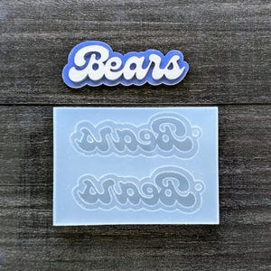 May include: A blue and white "Bears" word art design and a silicone mold with the word "Bears" in a retro font. The word art is in a blue outline with white lettering. The mold is light blue and has two impressions of the word.