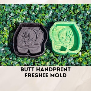 May include: Two silicone molds for making fresh-baked goods. The molds are shaped like a human bottom with a handprint on the back. One mold is black and the other is green. The text "BUTT HANDPRINT FRESHIE MOLD" is printed below the molds.
