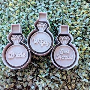 May include: Three gray plastic cookie cutters shaped like engagement rings with the words "Bride", "Wifey", and "Just Married" on them.