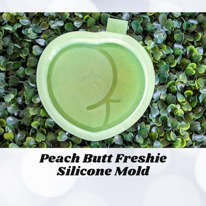 May include: A green silicone mold shaped like a peach with a peach pit. The mold is on a green leafy background. The text "Peach Butt Freshie Silicone Mold" is below the mold.