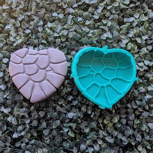 May include: Two heart-shaped molds. One is a light purple color with a textured stone pattern. The other is a bright teal color, also heart-shaped, with a similar stone pattern on the inside. The molds are likely for crafting or baking.