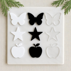 May include: A white square tile with nine decorative shapes: three butterflies, three stars, and three apples. The shapes alternate between white, black, and a clear outline. The tile is on a white surface, with green sprigs in the corners.
