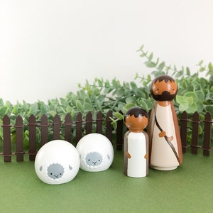 May include: Wooden peg dolls depicting a religious scene. Two white sheep with gray faces are in front of a brown picket fence. Two human figures with brown hair and robes stand behind the sheep. The background is green and white.