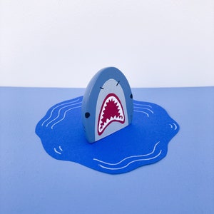 Könnte beinhalten: Ein Holzspielzeug in Form eines Hais. Der hellblaue Hai hat graue Details und ein rot-weißes Maul. Er taucht aus einem blauen Wasserausschnitt auf hellblauem Hintergrund auf. Das Spielzeug ist im Cartoon-Stil gestaltet.