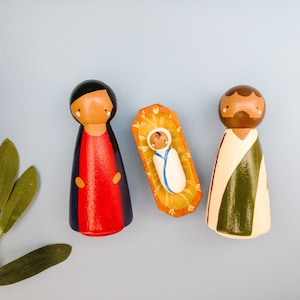 Op de afbeelding: Handgeschilderde houten kerststal met drie figuren: Maria in een rode en blauwe jurk, Jozef in een witte en groene mantel en baby Jezus in een kribbe. De achtergrond is lichtblauw.