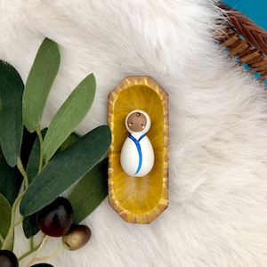 May include: A small, white ceramic figurine of a baby with brown eyes and a blue stripe on its white onesie. The figurine is sitting in a yellow wooden cradle.