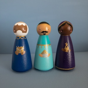 Puede incluir: Tres muñecas de madera pintadas a mano en azul, turquesa y morado. Cada muñeca tiene un diseño único de rostro y atuendo con detalles dorados. Las muñecas están de pie sobre un fondo azul claro.